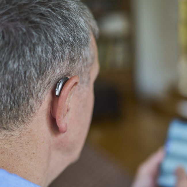 Close Up Of Mature Man Wearing Wireless Hearing Aid Using Mobile Phone At Home Close Up Of Mature Man Wearing Wireless Hearing Aid Using Mobile Phone At Home